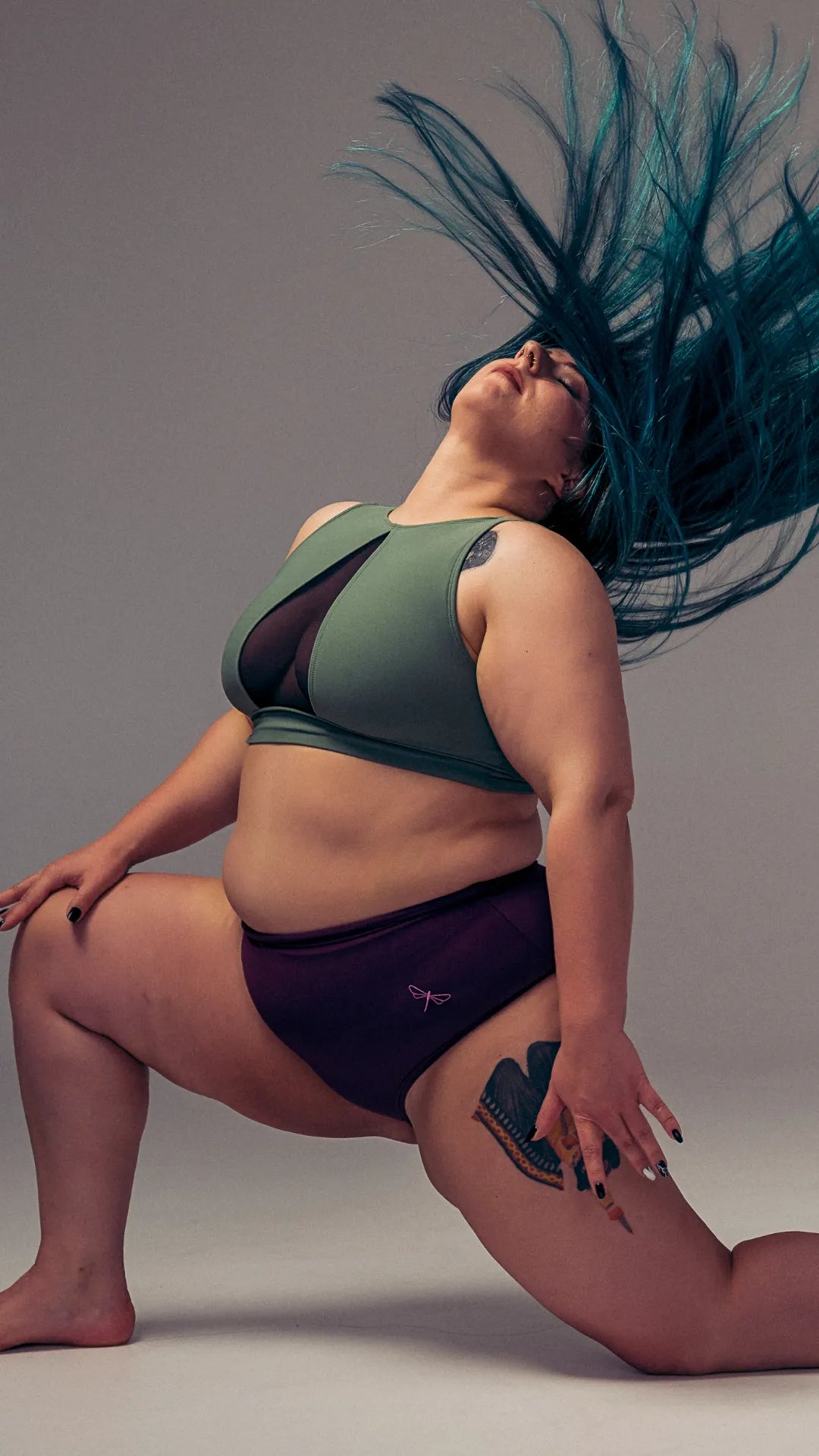 Kniende Tänzerin im salbeigrünen Mesh-Sport-BH und in pflaumenfarbenen Pole-Shorts im Hochformat